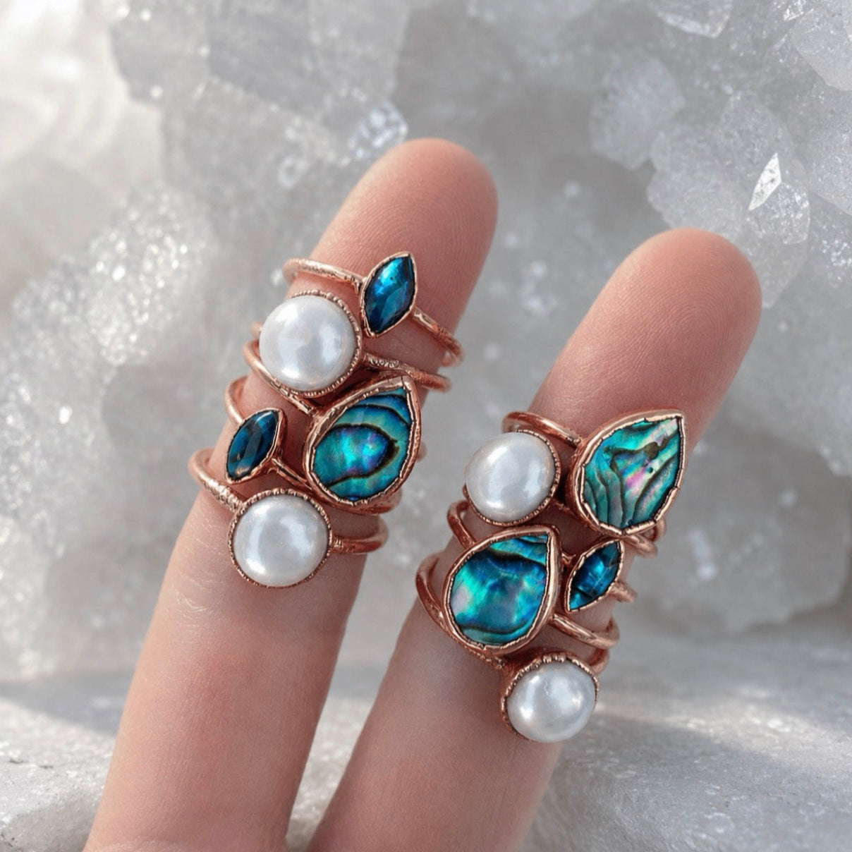 A selection of copper decorative rings with pearls and blue gemstones on a finger against a textured white background.