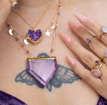 A close-up lifestyle shot of a person wearing a layered gold necklace with a large lavender-purple hexagonal crystal pendant, a smaller purple amethyst heart dizi pendant, and gold moon and star charms, alongside several gold rings on their hand.