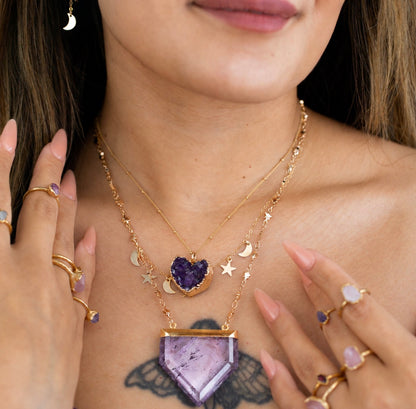 A close-up lifestyle shot of a model wearing a layered gold necklace set with a heart-shaped purple amethyst pendant, crescent moon charms, and star charms. The model also wears multiple gold rings with light purple gemstones.