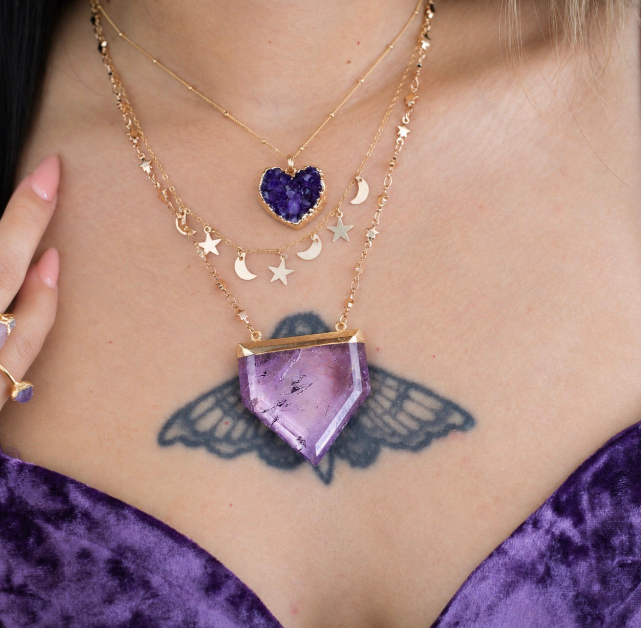 A close-up of a person wearing layered gold necklaces, including a large purple crystal pendant, smaller gold moon and star charms, and a purple heart-shaped pendant. A purple velvet top and a winged tattoo are also visible.
