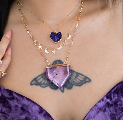 A close-up of a person wearing layered gold necklaces, including a large purple crystal pendant, smaller gold moon and star charms, and a purple heart-shaped pendant. A purple velvet top and a winged tattoo are also visible.