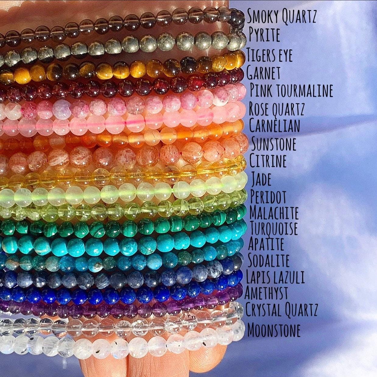 A hand holding 19 strands of different gemstones, in color order to match a rainbow. Beads are 4mm in size and polished, with a round shape. You get topick which crystal calls to you.